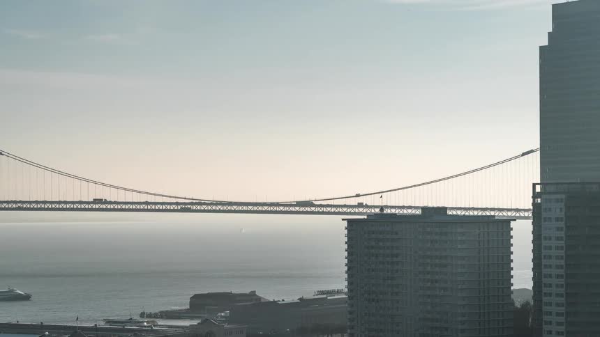 A hazy cityscape shot features a suspension bridge spanning across a body of water partially obscured by tall buildings in the foreground