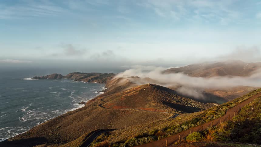 An aerial view shows a winding road traversing a hilly coastal landscape with the ocean on one side and rolling hills on the other under a partly cloudy sky