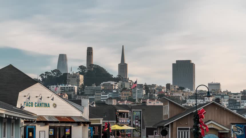 The san francisco skyline featuring coit tower and the transamerica pyramid rises behind waterfront shops under a cloudy sky