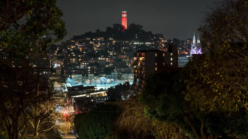 A night timelapse from a hillside captures city traffic streaks below with buildings and the iconic redlit coit tower standing out in the distance