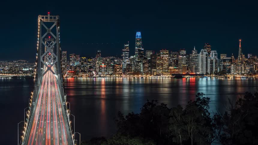 A bunch of cars are going across the bridge in the night time and the skyline is lit up by the skyscrapers