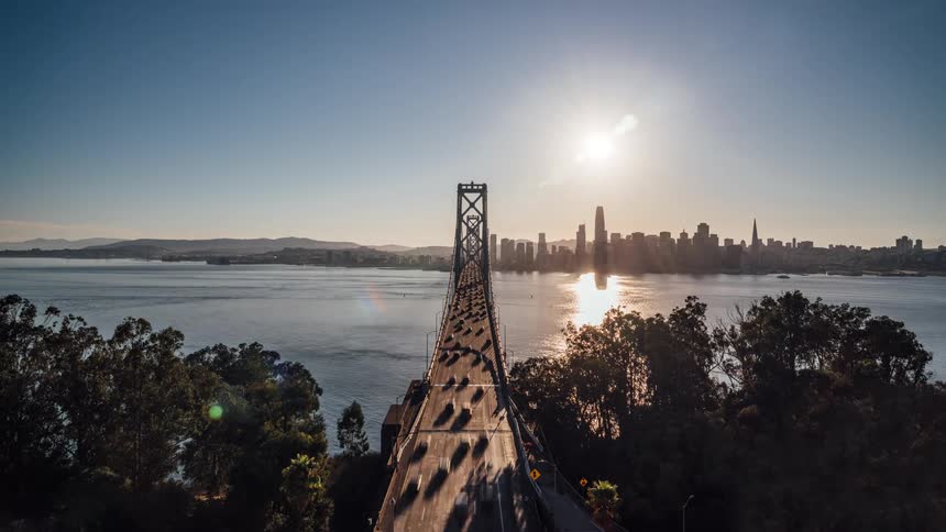 Streaks of traffic blur across a long bridge in this timelapse facing a city skyline all set against the setting sun