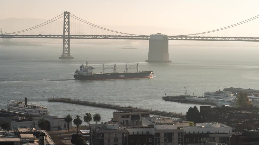 A large cargo ship sails under the bay bridge with a pier and city buildings visible in the foreground