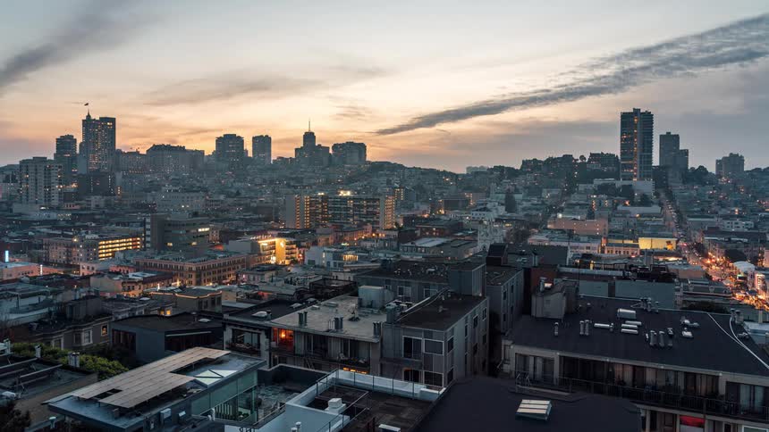 A static camera captures the sun moving from dawn to dusk and into the night over a bustling metropolis with clouds moving fluidly above tall buildings and lights blurring during the night through the city