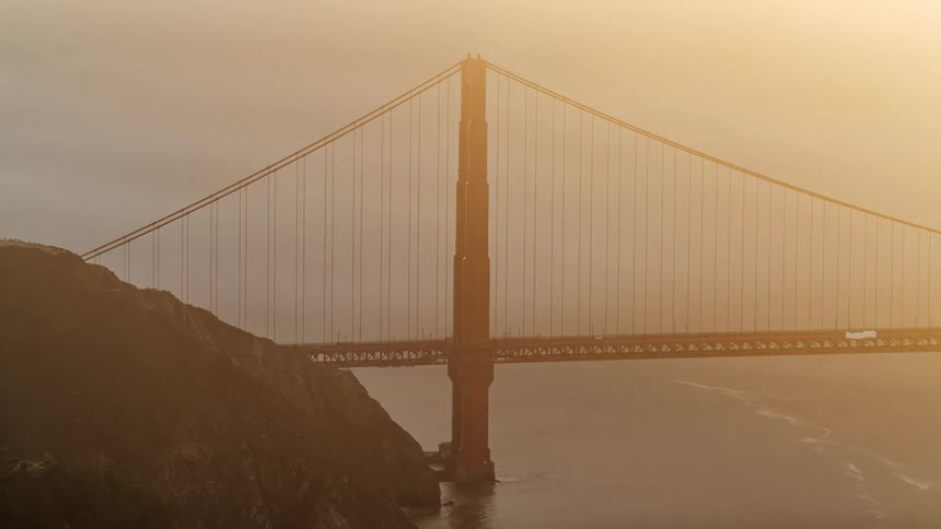 The golden gate bridges tower and cables partially obscured by a hill is bathed in a warm golden light