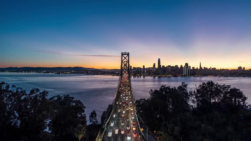 An aerial view shows cars crossing the bay bridge as it stretches across the water towards the san francisco skyline at sunset with trees in the foreground