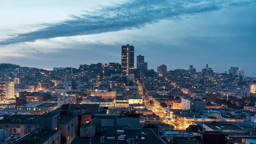 A cityscape view shows buildings nestled on rolling hills gradually illuminated as the day ends under a soft sunrise sky