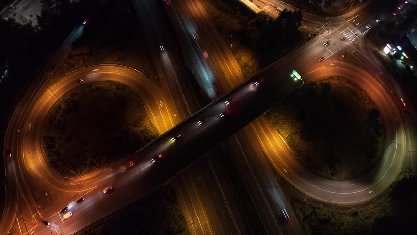 Aerial view of a highway interchange at night with streaks of red and white lights from moving vehicles creating a dynamic contrast against the dark roads and warm street lighting