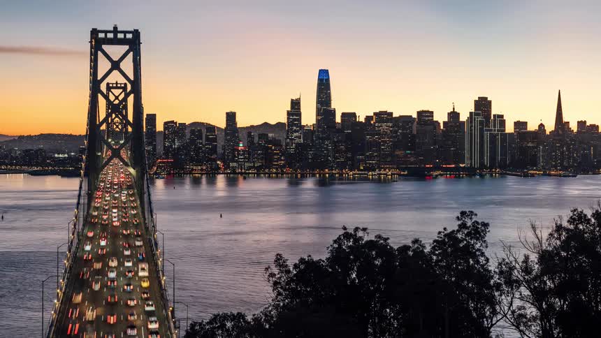 An aerial view shows cars crossing the bay bridge as it stretches across the water towards the san francisco skyline at sunset with trees in the foreground