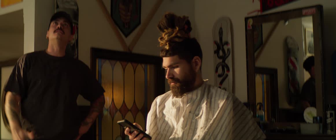 A man in a barber cape looks at his phone while another man has a conversation nearby