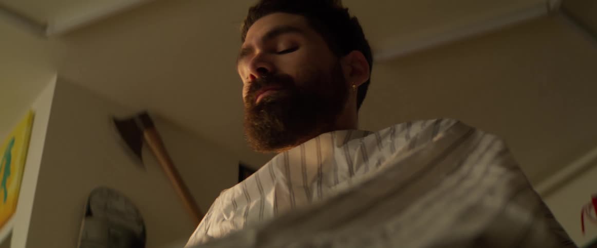 A bearded man sits in a barbershop with a coat draped over him waiting