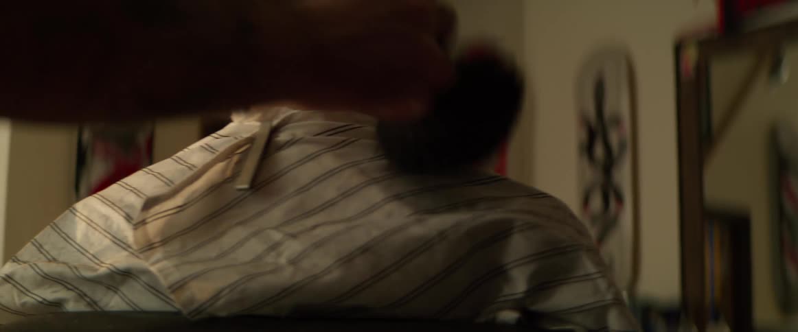 A barber brushes the shoulders of a seated customer wearing a cape in a dimly lit room
