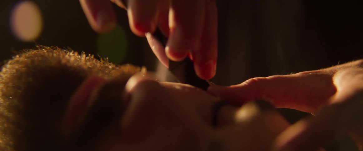 Closeup of a mans face as he lies down while a barber trims his beard with a razor blade in a dimly lit room
