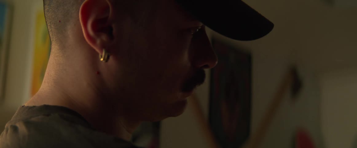 A closeup of a mans head wearing a baseball hat with an earring and a mustache in a dimly lit room looking at something