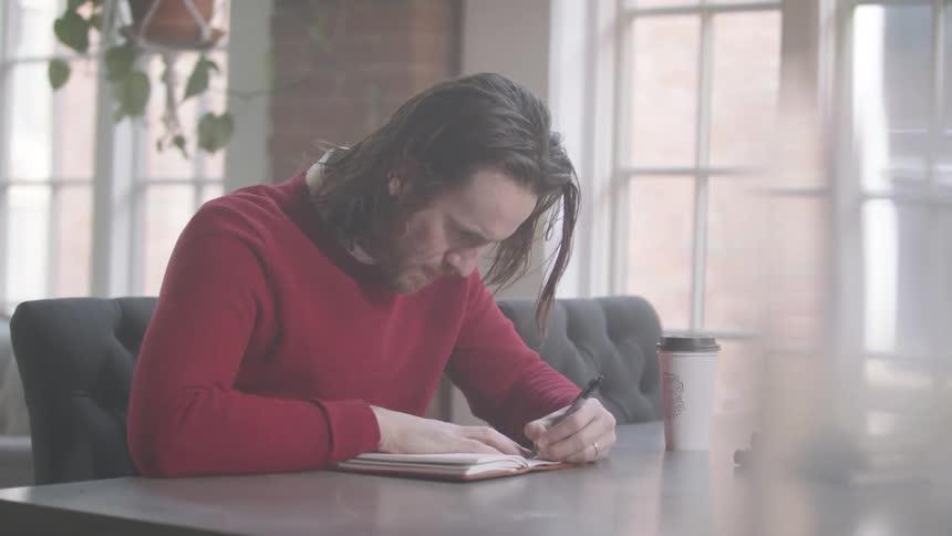 A man with long dark hair is writing in a notebook at a table in a bright room with large windows