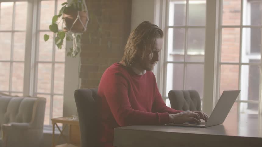 Man sitting at a table near windows typing on a laptop computer