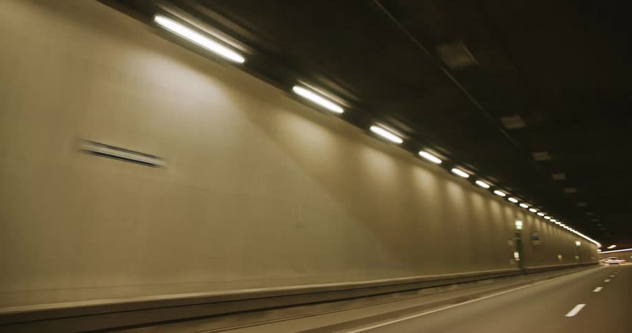 The camera pans from the lights alongside a road tunnel wall to the road itself which is empty of other vehicles and clean showing an uninterrupted row of lights along the tunnel and evenly spaced road markings on the highway