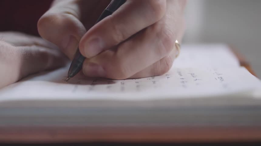 In a tight closeup a hand wearing a simple ring carefully guides a pen across the lined paper of an open notebook diligently writing