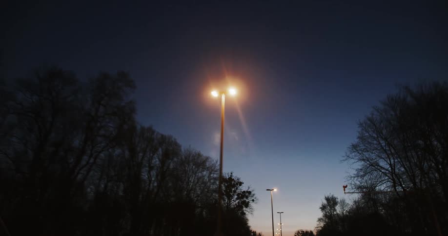 Several street lamps illuminate a road at dusk with a car driving in the distance
