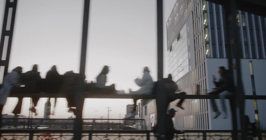 A lot of people are sitting on the raiiing or walking on a bridge in a city