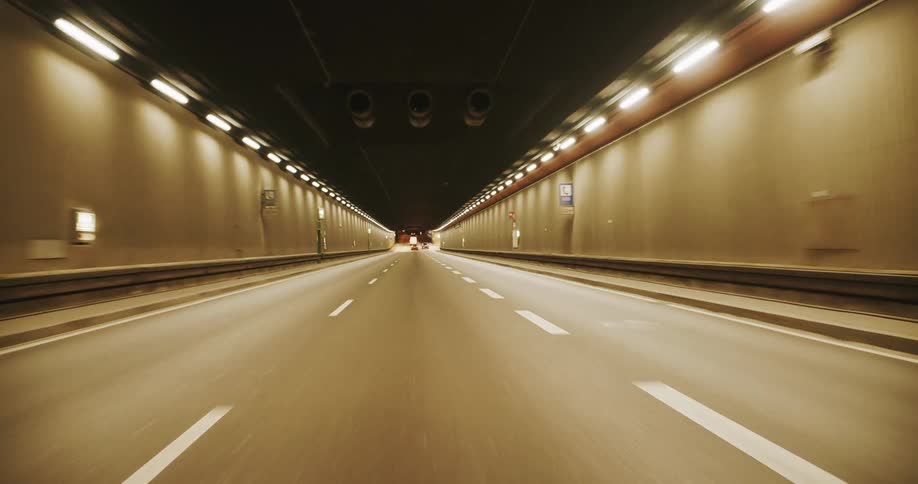 The perspective moves steadily through a brightly lit roadway tunnel at night where rows of overhead and wallmounted lights illuminate the path ahead revealing lane markings and distant taillights