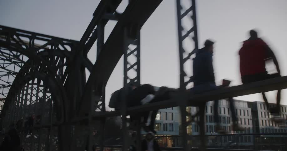 People sitting on a cross piece of the side of a bridge and leaning on the rail on the nearby walkway look at the water and the city