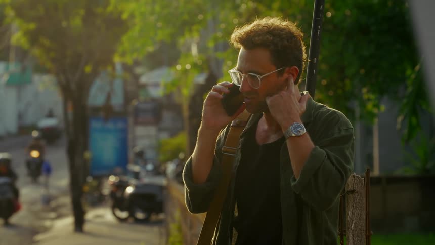 A man with curly hair and glasses illuminated by the setting sun is engrossed in a phone call on a city street lined with trees and parked motorcycles