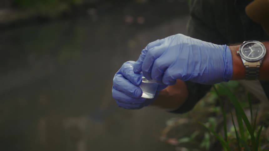 A person stands beside a muddy drainage canal wearing blue gloves and collecting a water sample in a small bottle with a lid