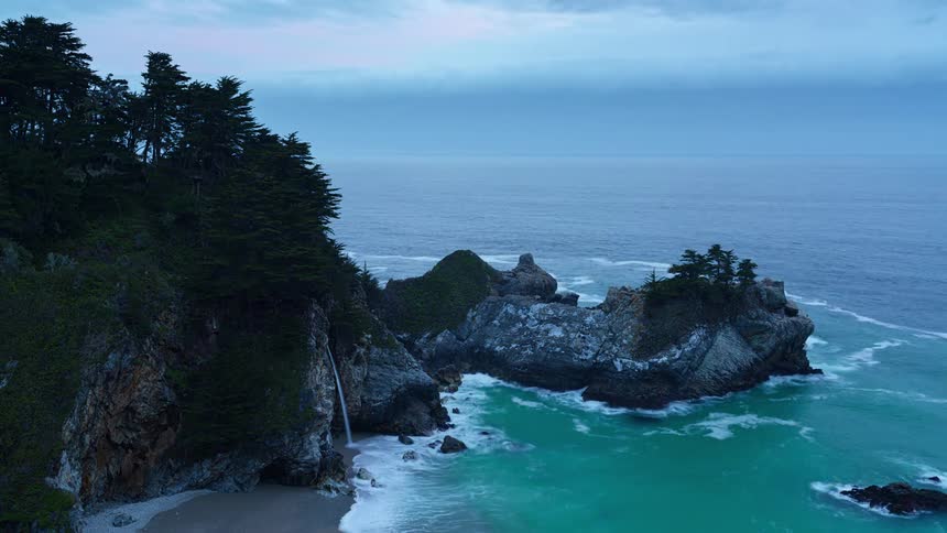 A twilight aerial view captures a waterfall cascading onto a secluded beach cove surrounded by rocky cliffs and the vast ocean