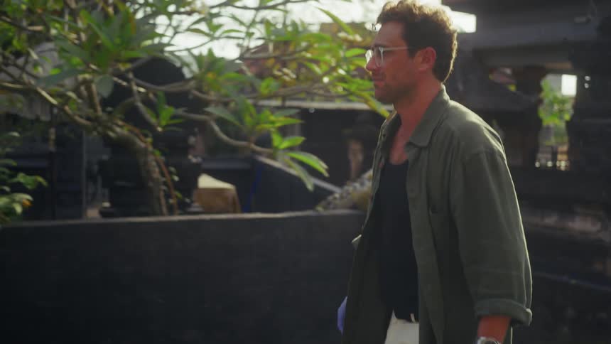 A man with glasses and a green shirt walks past traditional balinese structures in bright sunlight