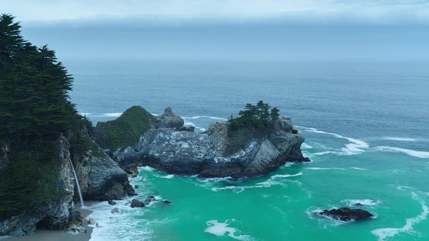 An aerial view of a waterfall cascading onto a sandy beach cove with turquoise waters surrounded by rocky cliffs and lush vegetation under an overcast sky