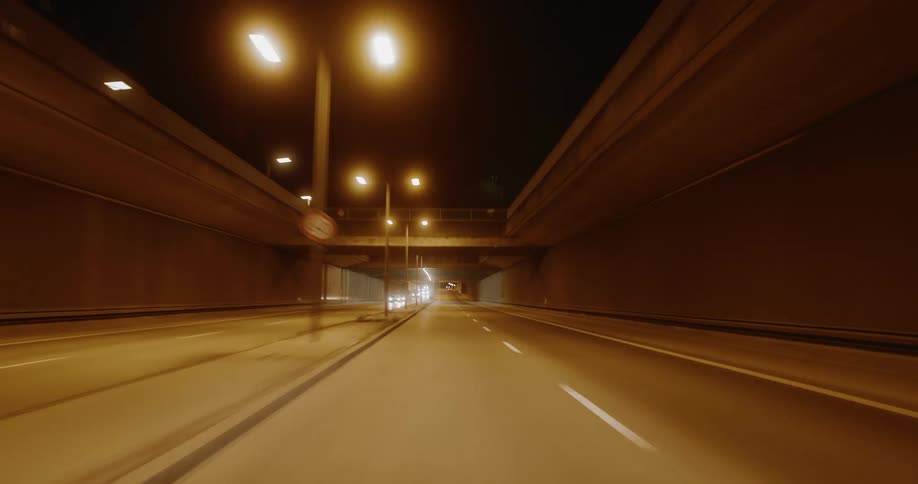 From a driving pov on a multilane highway at night concrete retaining walls bathed in the warm glow of overhead street lamps line the road as faint headlight streaks from distant oncoming traffic cut through the darkness