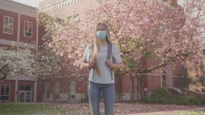 Blonde female student wearing face mask standing in front of cherry blossom tree at university