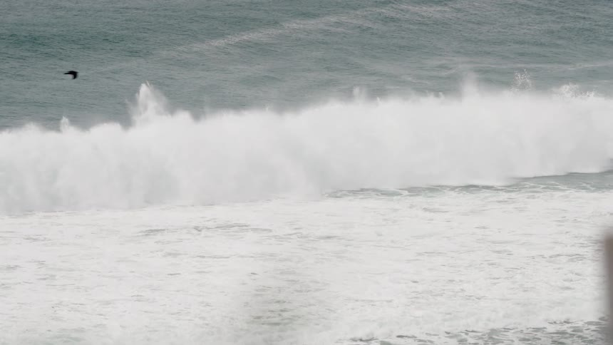 A massive ocean wave with a turquoise face curls powerfully as it crests and begins to break sending plumes of white spray into the air above the churning sea surface under an overcast sky