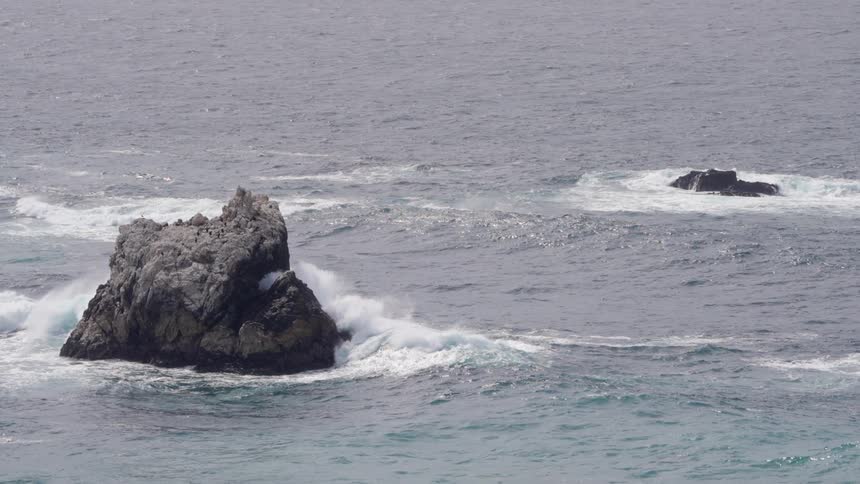 Turbulent ocean waves crash vigorously against a large dark craggy rock formation rising from the choppy grey sea generating significant white water foam and spray under diffused overcast lighting conditions