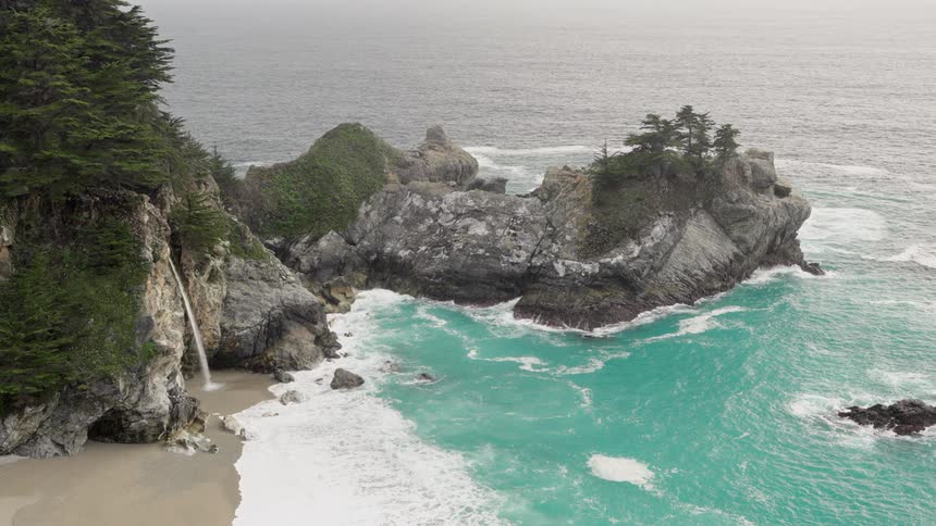 A waterfall flows onto a sandy beach while ocean waves crash against a nearby promontory