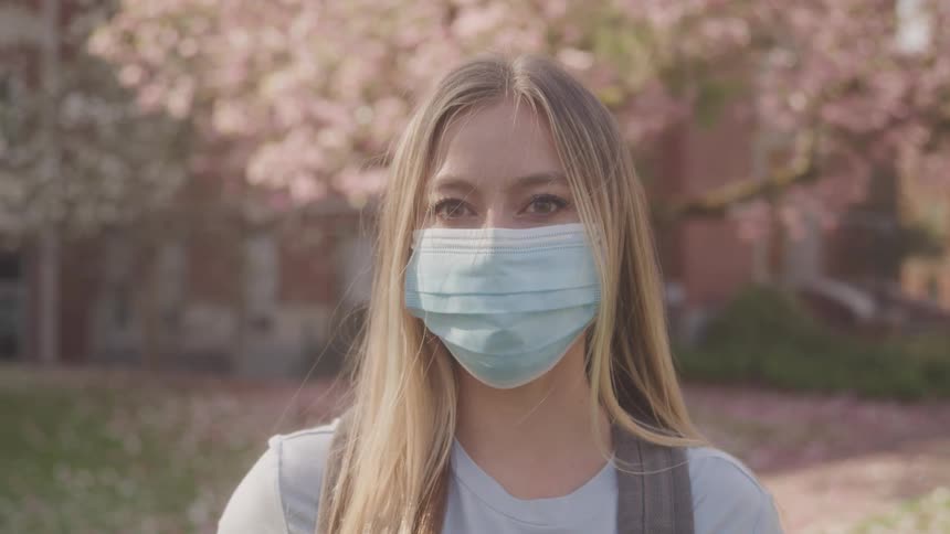 Against a softfocus background of spring blossoms and brick a young woman possibly a student with her backpack wears a blue face mask in this outdoor portrait