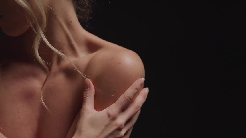 A shirtless woman with blonde hair focusing on her shoulder and hand against a dark background where her hand gently rests on her shoulder