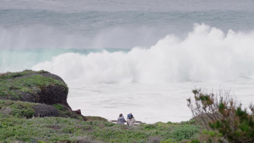 Viewed from an elevated hilltop behind them three hikers observe a spectacular large wave crashing onto the rocky coast showcasing the raw power of the ocean