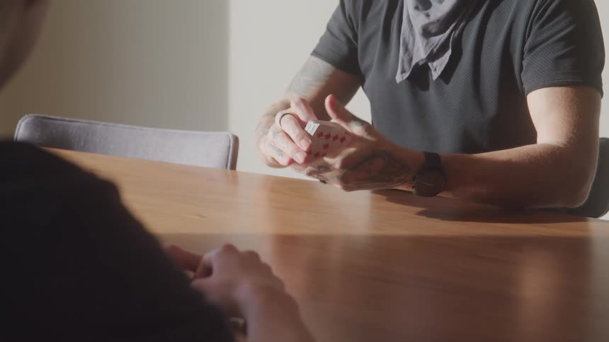 Man shuffles cards at the table in preparation for a game while another person waits to begin