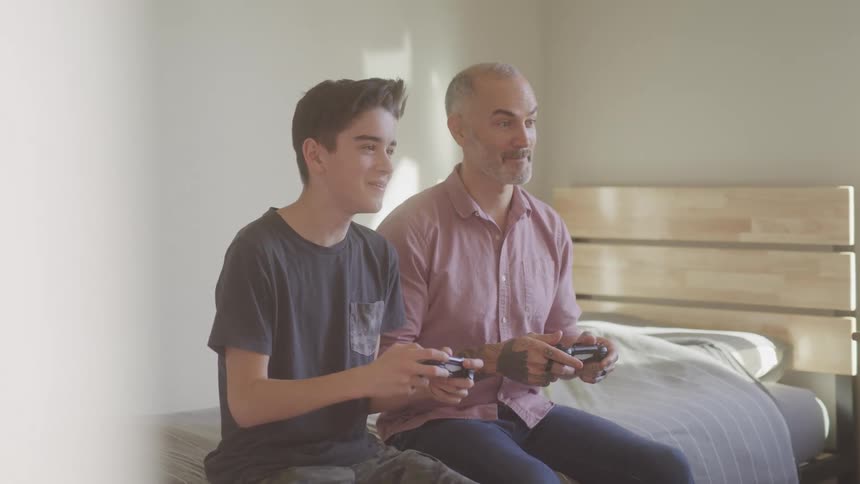 A father and son are sitting on the bed playing video games