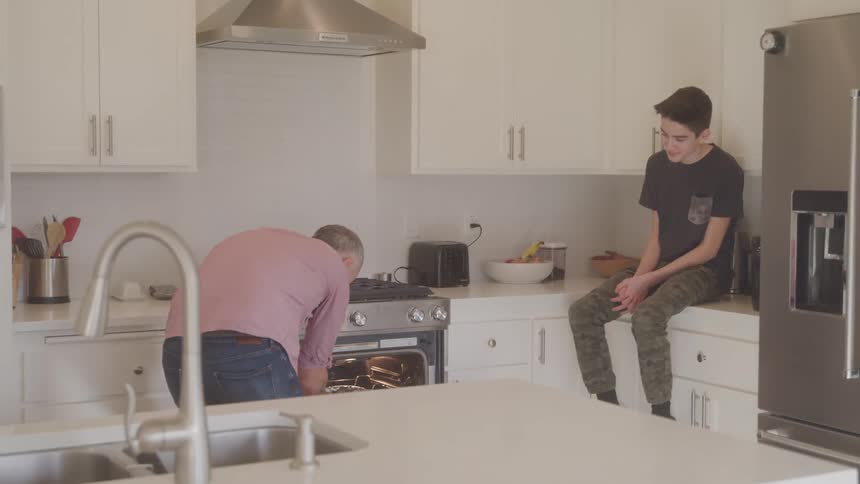A teenage boy watches his dad pull food out of the oven