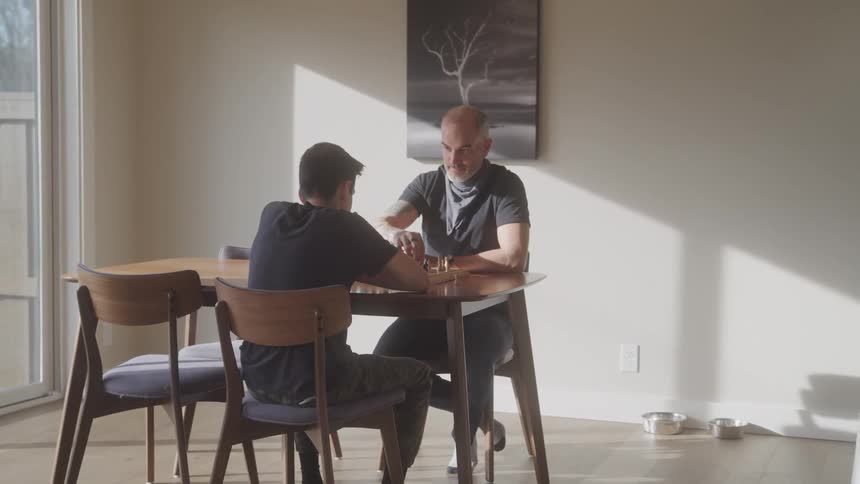 Two men sitting around a wooden table and playing chess near a window