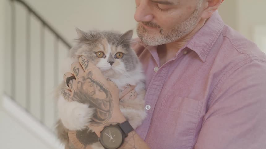 A man with a salt and pepper beard and a pink shirt holds a fluffy gray and white cat in his arms
