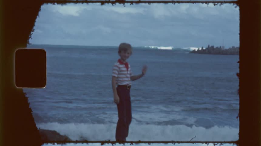 A young boy waves while standing in shallow ocean water at sunset