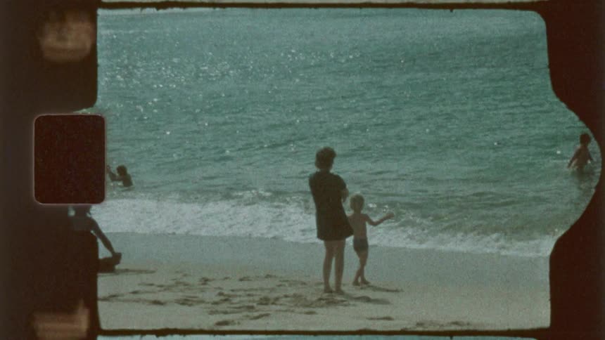 A grainy film shows a beach scene with waves reaching the shore as a mother and child stand facing the ocean