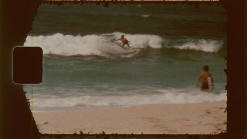 A vintage film shows a surfer riding a wave followed by another person walking into the ocean with a surfboard