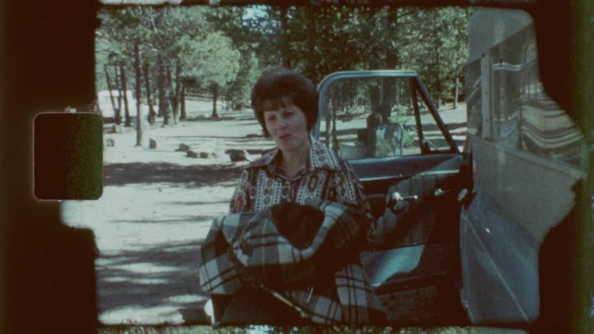 Woman carrying a coat and a purse smiles while walking away from the open door of a vehicle parked near the woods