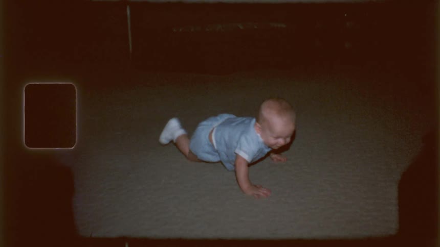 A baby in a light blue romper crawling on a dark carpet with a grainy texture