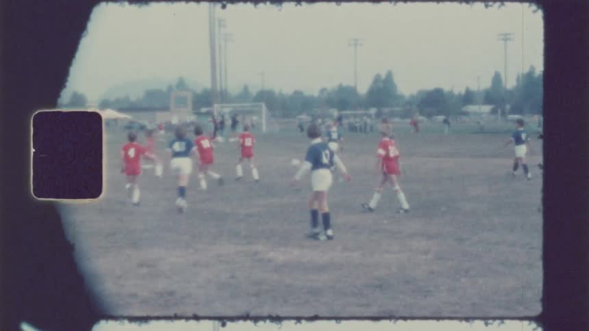 A youth soccer game in progress on a grassy field with players in red and blue jerseys running towards the goal while spectators and tents are visible in the blurred background under an overcast sky